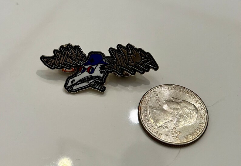 Puede incluir: Un pin de esmalte plateado con un cr&aacute;neo de dibujos animados que lleva una gorra de b&eacute;isbol azul y gafas de sol. El cr&aacute;neo tiene un dise&ntilde;o de rayo en cada lado de su cabeza. El pin es aproximadamente del tama&ntilde;o de una moneda de 20 c&eacute;ntimos de euro.