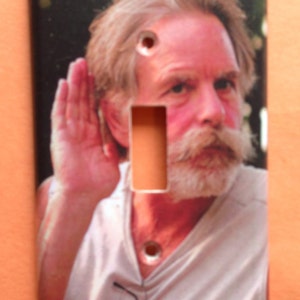 May include: Rectangular light switch cover featuring a man with a white mustache and beard, with his hand cupped to his ear. The cover has two screw holes and a rectangular opening for the switch.