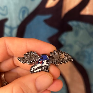 Puede incluir: Un pin de metal plateado con un alce de dibujos animados que lleva un sombrero azul y gafas de sol. El alce tiene un dise&ntilde;o de rayo en sus astas.