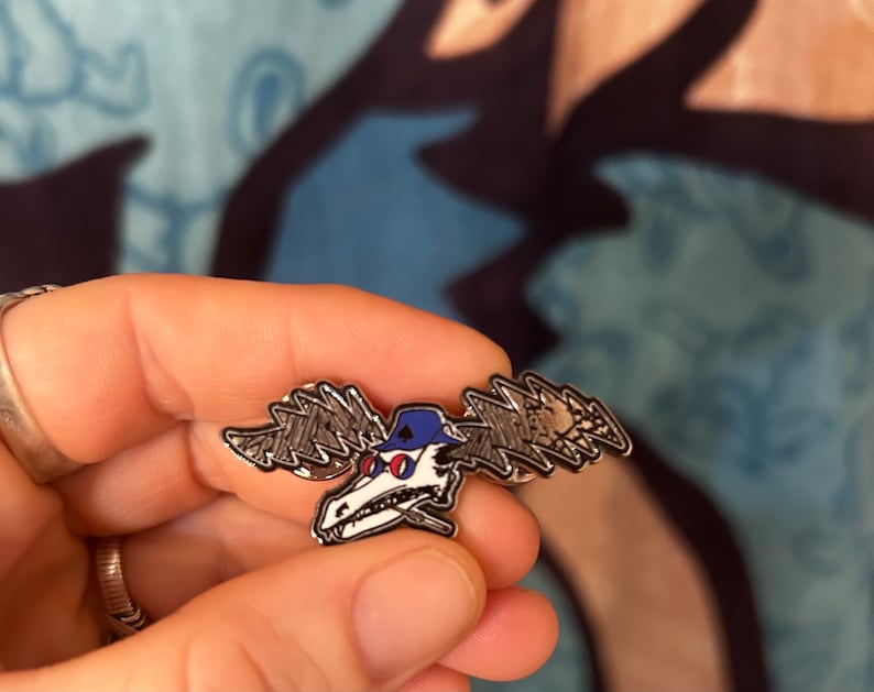 Puede incluir: Un pin de metal plateado con un alce de dibujos animados que lleva un sombrero azul y gafas de sol. El alce tiene un dise&ntilde;o de rayo en sus astas.