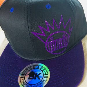 May include: Black and purple snapback hat with a flat brim. The hat features a purple glitter crown design and the word "Kings" in purple glitter. The hat has a "BK Caps" sticker on the brim.