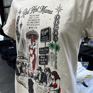 May include: A light beige t-shirt with a graphic design featuring the words "Red Hot Mama" and a Las Vegas theme. The design includes illustrations of showgirls, casinos, and the text "May 8,9,&10 2026."