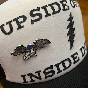 Puede incluir: Gorra de camionero blanca con texto negro de purpurina que dice "UPSIDE OUT INSIDE DOWN". La gorra tambi&eacute;n tiene un rayo negro de purpurina y un pin de alce plateado.