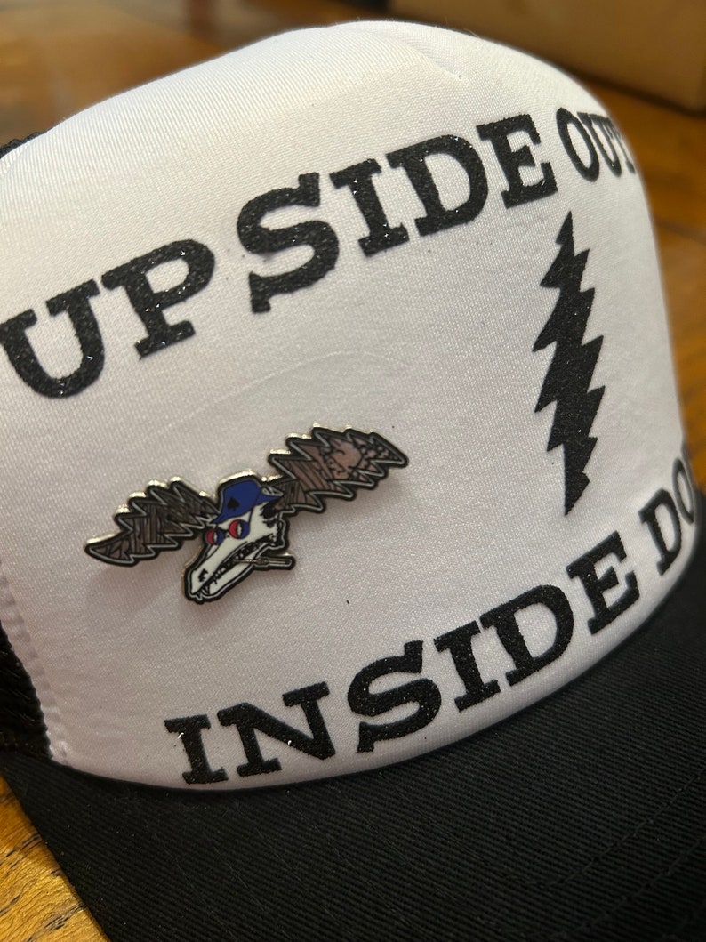 Puede incluir: Gorra de camionero blanca con texto negro de purpurina que dice "UPSIDE OUT INSIDE DOWN". La gorra tambi&eacute;n tiene un rayo negro de purpurina y un pin de alce plateado.