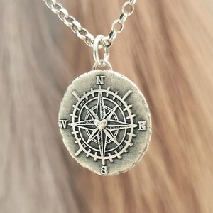 Może przedstawiać: Srebrny naszyjnik z zawieszką w kształcie kompasu. Kompas ma szczegółowy wzór z sercem w środku.