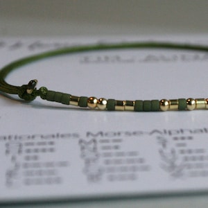 Könnte beinhalten: Ein grünes und goldenes Perlenarmband mit einem verschiebbaren Knotenverschluss. Das Armband besteht aus kleinen, runden Perlen und liegt auf einem weißen Hintergrund.