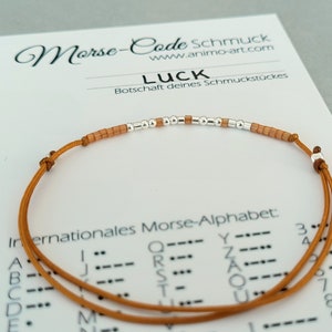 May include: A brown cord bracelet with silver and orange beads spelling out the word "LUCK" in Morse code. The bracelet is on a white card with the text "Morse-Code Schmuck www.animo-art.com LUCK Botschaft deines Schmuckstückes Internationales Morse-Alphabet:"