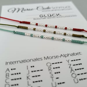 May include: Three bracelets made with silver, green, and red beads. The bracelets are on a white card with the word "GLÜCK" and the text "Botschaft deines Schmuckstückes" in German. The card also includes a chart of the international Morse code alphabet.