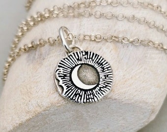 May include: A silver-toned necklace featuring a circular pendant with a crescent moon design at the center, surrounded by radiating etched lines. The pendant hangs from a delicate, thin silver-toned chain with small, round, interconnected links.