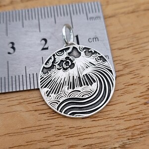 Könnte beinhalten: Ein silberner Anhänger mit einem detaillierten Schwarz-Weiß-Design einer Welle, die gegen Wolken kracht. Der Anhänger hat einen Durchmesser von etwa 1 cm.