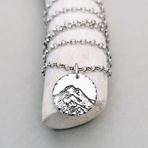 May include: A silver necklace with a round pendant featuring a detailed engraved mountain scene. The pendant has a textured edge.