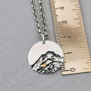 May include: Silver necklace with a round pendant featuring a black and white mountain scene with a small gold heart. The necklace is hanging from a ruler.