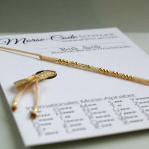 Puede incluir: Un brazalete de cuentas blancas y doradas con el mensaje "BIG SIS" en código Morse. El brazalete está sobre una tarjeta blanca con el texto "Morse-Code Schmuck" y "Botschaft deines Schmuckstückes".