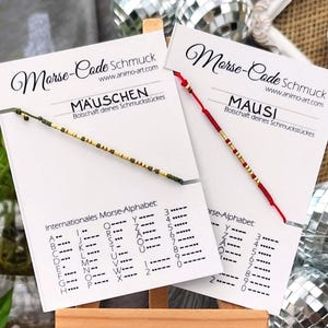 Könnte beinhalten: Zwei Morsecode-Armbänder auf weißen Präsentationskarten. Ein Armband hat eine olivgrüne Schnur mit Goldperlen und ist mit MAUSCHEN beschriftet. Das andere hat eine rote Schnur mit Goldperlen und ist mit MAUSI beschriftet. Internationales Morse-Alphabet.