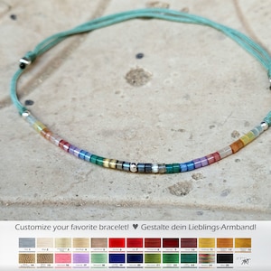 Könnte beinhalten: Ein türkisfarbenes Kordelarmband mit einer Reihe bunter Perlen, darunter grün, blau, lila, rot, orange und gelb. Das Armband ist verstellbar und hat silberne Metallenden. Das Bild zeigt auch eine Farbkarte mit verschiedenen Kordelfarben.