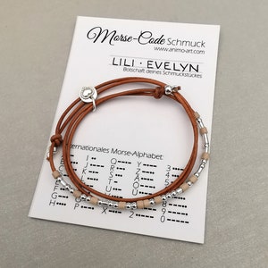 Könnte beinhalten: Ein braunes Lederarmband mit silbernen und beigefarbenen Perlen. Das Armband ist um eine Karte mit dem Text "Morse-Code Schmuck" und "LILI EVELYN" in schwarzer Schrift gewickelt. Die Karte enthält auch eine Morse-Code-Alphabet-Tabelle.