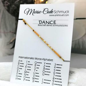 Pode incluir: Uma pulseira de cordão amarelo com pequenas contas amarelas e douradas dispostas num padrão de código Morse. A pulseira está presa a um cartão branco com a palavra DANCE e uma tabela do alfabeto morse internacional.
