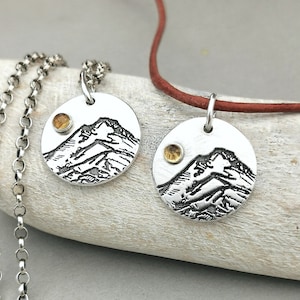 May include: Two silver necklaces with round pendants featuring a mountain scene and a small yellow gemstone. One necklace has a silver chain and the other has a brown leather cord.