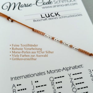 Könnte beinhalten: Ein braunes und silbernes Perlenarmband mit dem Wort "LUCK" auf einer Karte. Die Karte enthält auch eine Beschreibung der Eigenschaften des Armbands, wie z. B. feine Textilbänder, robuste Verarbeitung und 925er Silberperlen. Die Karte enthält auch ein internationales Morse-Alphabet.