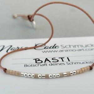 Puede incluir: Un brazalete de cordón marrón con cuentas plateadas y beige que deletrean el nombre "BAsTI". El brazalete está sobre una tarjeta blanca con el texto "Code Schmuck www.animo-art.com" y "Botschaft deines Schmuckstil".