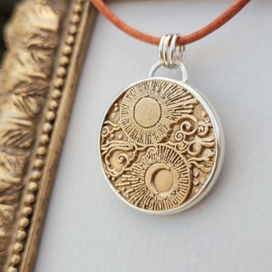 May include: A silver necklace with a round pendant featuring a gold sun and moon design with clouds. The pendant has a detailed, textured surface.