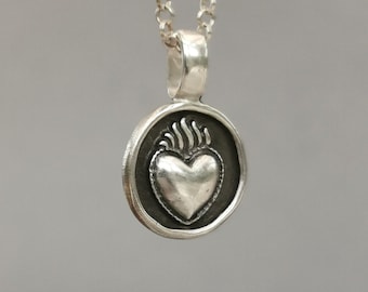 Op de afbeelding: Zilveren ketting met een ronde hanger met een gestileerd hart met vlammen. De hanger is zwart en zilver.