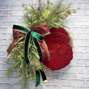 May include: A festive Christmas wreath featuring a red Santa Claus face, adorned with a plaid and green velvet bow. The wreath is decorated with pine needles, sprigs, and small pine cones, perfect for holiday decor.