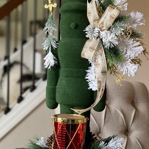 May include: A green felt nutcracker figurine wearing a gold crown and holding a gold fleur-de-lis scepter. The nutcracker is standing on a red drum with gold trim. The nutcracker is surrounded by a white and green Christmas wreath.