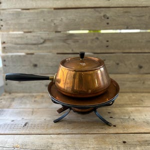May include: A copper fondue pot with a black handle and a matching lid featuring a black knob. The pot rests on a circular copper plate supported by a black metal stand with four curved legs, designed for heating and serving food at the table.