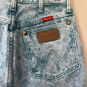 May include: Acid-wash light blue denim jeans featuring a high-waisted design with belt loops. A brown leather patch with the Wrangler logo is stitched onto the back pocket, which also displays a signature W-shaped stitch pattern and copper rivets.