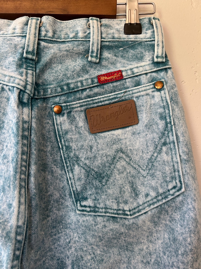 May include: Acid-wash light blue denim jeans featuring a high-waisted design with belt loops. A brown leather patch with the Wrangler logo is stitched onto the back pocket, which also displays a signature W-shaped stitch pattern and copper rivets.