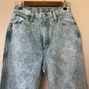 May include: High-waisted denim jeans featuring a light blue acid wash finish. The pants include a classic five-pocket design with copper-toned metal rivets at the pocket corners, a button fly closure, and visible stitching along the seams.