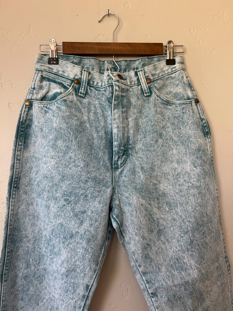 May include: High-waisted denim jeans featuring a light blue acid wash finish. The pants include a classic five-pocket design with copper-toned metal rivets at the pocket corners, a button fly closure, and visible stitching along the seams.