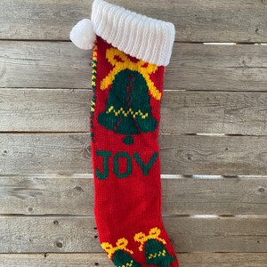 May include: Red knitted Christmas stocking with a white cuff and pom pom. The stocking features green and yellow bells with bows and the word "JOY" in green letters.