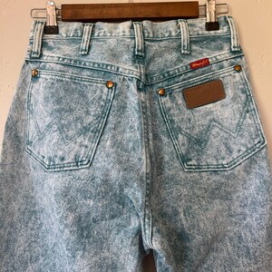 May include: Light blue acid-wash denim jeans featuring a high-waisted design, two back pockets with a signature W-stitch pattern, copper-toned rivets, and a brown leather Wrangler brand patch on the right pocket with a small red logo tag.