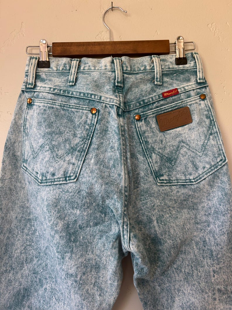 May include: Light blue acid-wash denim jeans featuring a high-waisted design, two back pockets with a signature W-stitch pattern, copper-toned rivets, and a brown leather Wrangler brand patch on the right pocket with a small red logo tag.