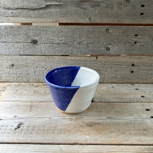 May include: A small, white ceramic bowl with a blue glaze on one half. The bowl is sitting on a wooden surface.