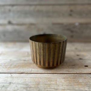 May include: A small, cylindrical brass bowl with vertical ribbed detailing. The bowl has a slightly flared base and a smooth interior. The brass has a warm, aged patina, giving it a vintage look. The bowl is empty and sits on a wooden surface.