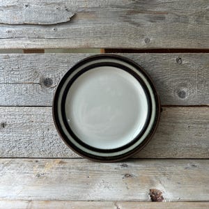 May include: A round ceramic plate with a cream-colored center and concentric brown and black bands around the rim. The plate is set against a weathered wooden background, showcasing its simple, yet elegant design. Ideal for serving meals.