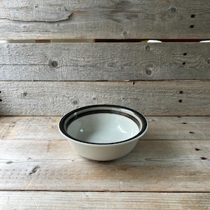 May include: A white ceramic bowl with a black and brown striped rim. The bowl is set on a weathered wooden surface. The bowl is empty and appears to be in good condition.