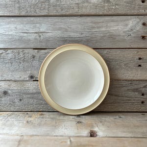 May include: A round, off-white ceramic plate with a tan rim. The plate is set against a rustic, weathered wooden background, creating a simple, neutral aesthetic. The plate is likely for serving food.