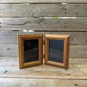 May include: A hinged double picture frame made of light brown wood with a visible grain pattern. The frame consists of two rectangular sections connected by metal hinges, each featuring a dark, empty interior space for displaying photographs.