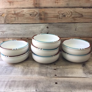 May include: A stack of six white ceramic bowls with a brown rim, a light blue stripe, and small blue dashes. The bowls are arranged on a weathered wooden surface, showcasing a rustic aesthetic. The bowls are empty and ready for use.