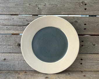 May include: A white ceramic plate with a wide rim and a dark gray center. The plate is sitting on a wooden surface.