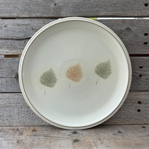 May include: A large, cream-colored ceramic dinner plate with a brown rim and three stylized leaf designs in shades of green and brown.