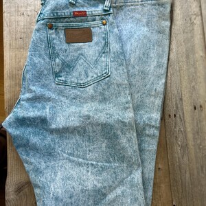 May include: Light blue acid-wash denim jeans featuring a high-waisted cut, a classic five-pocket design, and a brown leather patch on the back pocket with the Wrangler logo. The jeans include copper-colored rivets and a red Wrangler brand tag.
