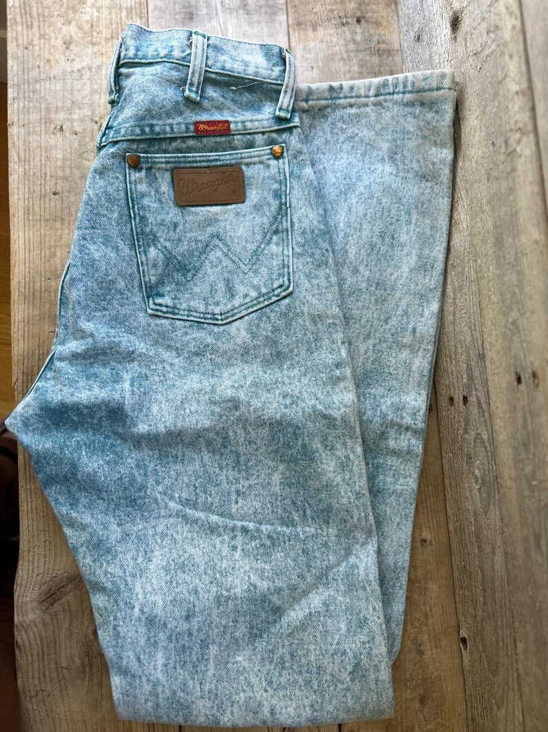 May include: Light blue acid-wash denim jeans featuring a high-waisted cut, a classic five-pocket design, and a brown leather patch on the back pocket with the Wrangler logo. The jeans include copper-colored rivets and a red Wrangler brand tag.
