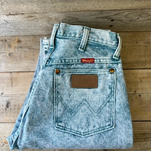 May include: Light blue acid-wash denim jeans featuring a high-waisted design with belt loops. A rectangular brown leather patch with the embossed Wrangler logo is centered on the back pocket, which also displays a stitched W pattern.