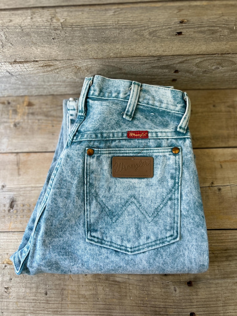 May include: Light blue acid-wash denim jeans featuring a high-waisted design with belt loops. A rectangular brown leather patch with the embossed Wrangler logo is centered on the back pocket, which also displays a stitched W pattern.