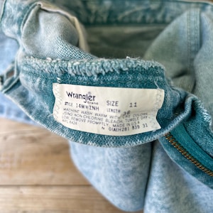 May include: Light blue acid-wash denim jeans featuring a white interior tag with the Wrangler brand logo, style number 14MWZNH, size 11, and length 36. The tag includes care instructions for machine washing and tumble drying, and states Made in USA.